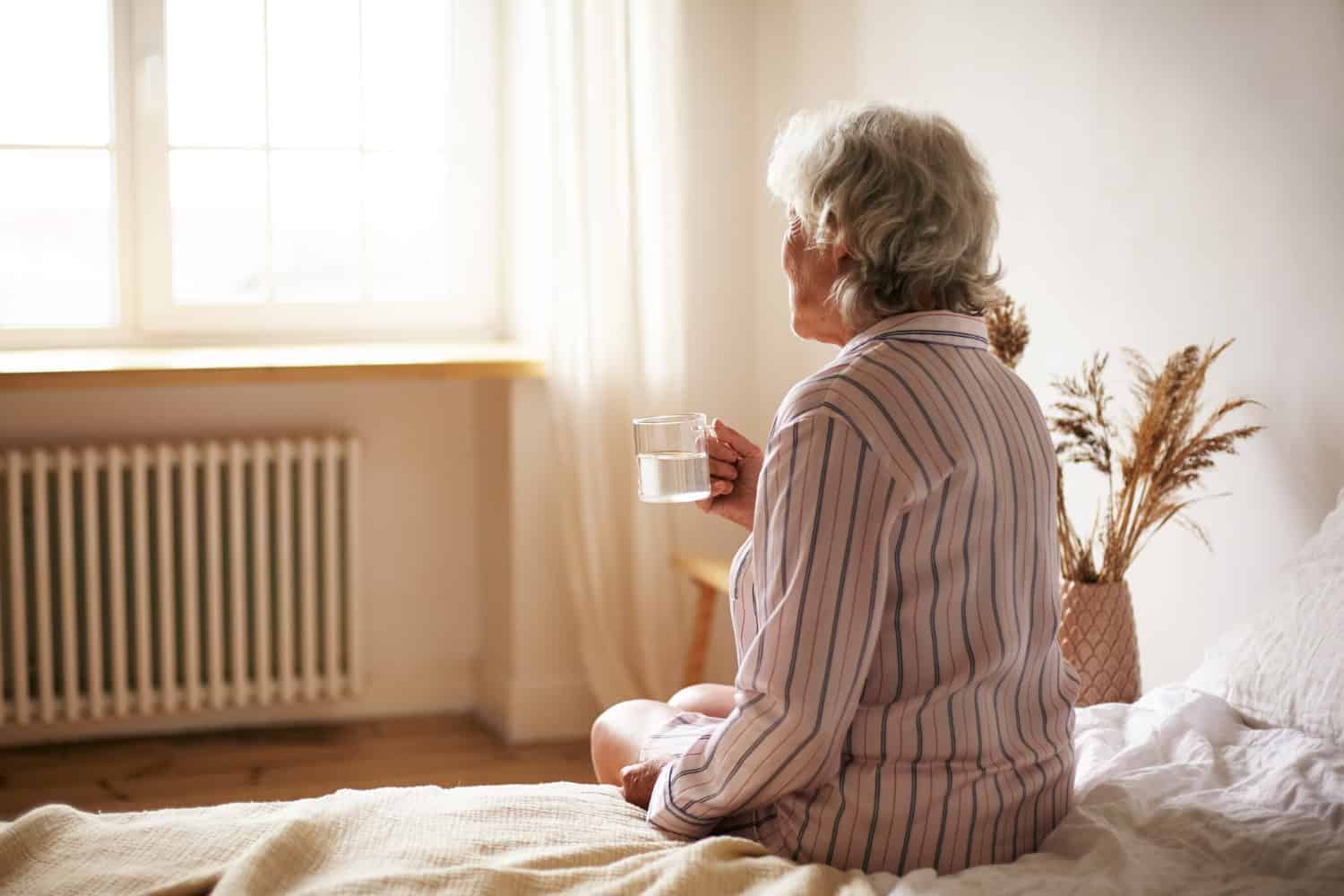 Picture of an older woman sitting on the edge of the bed, looking out the window, and holding a clear glass mug full of water.