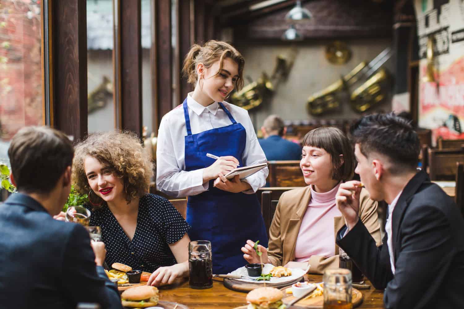 A waitress stands at a table of four seated restaurant guests while taking food orders.