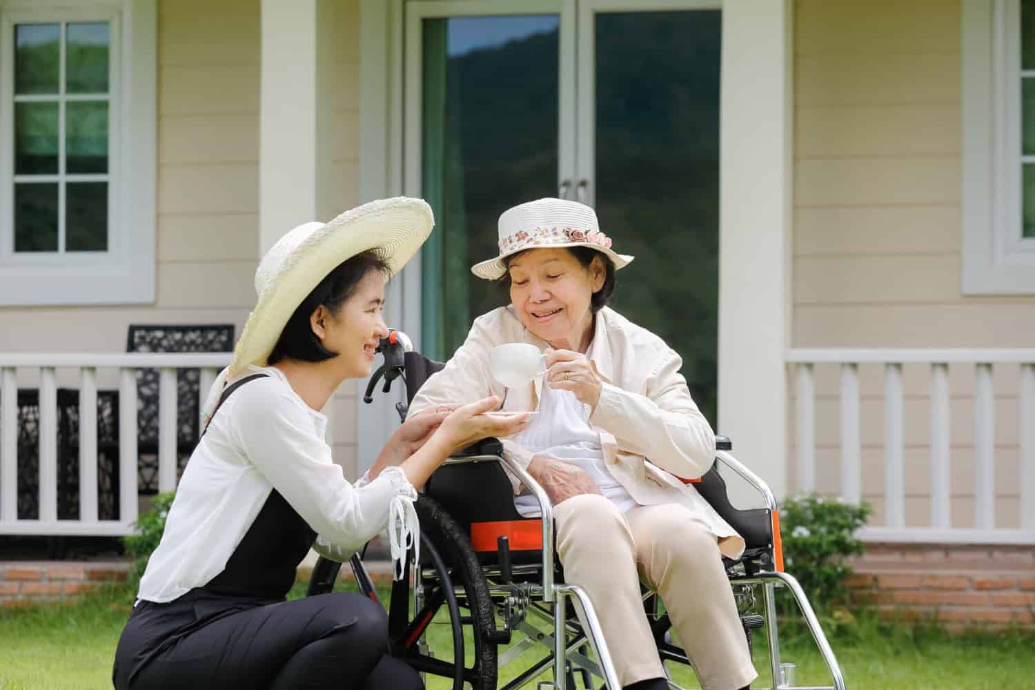 An elderly woman and her adult daughter sit outside, sharing a cup of tea and smiling at each other.