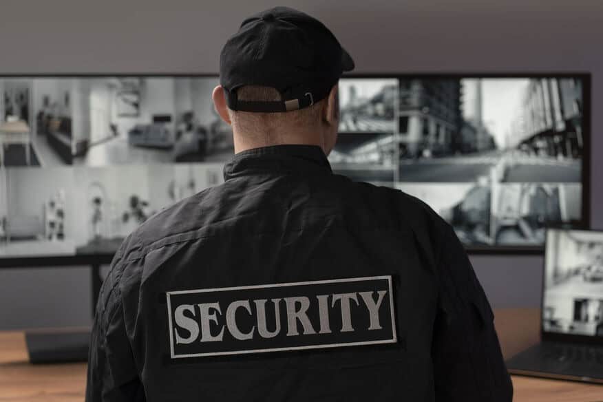 The back of man in a security jacket standing in front of a bank of monitors showing various black and white scenes. 