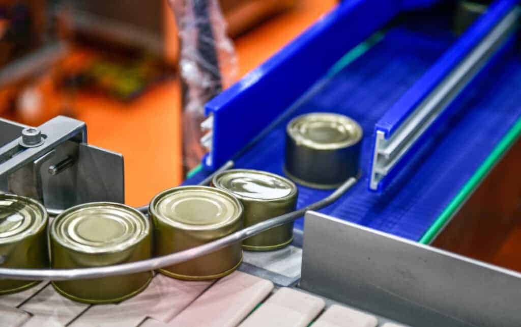 Metal cans move down a blue and silver metal conveyor belt in a food production facility.