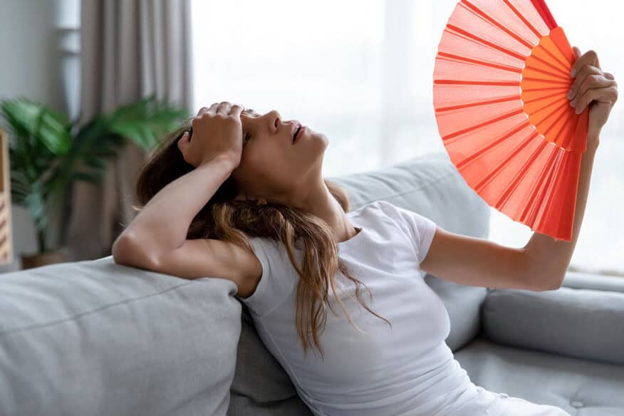 A woman is sitting on a couch indoors, fanning herself and showing discomfort in the humidity from poor hotel air quality.