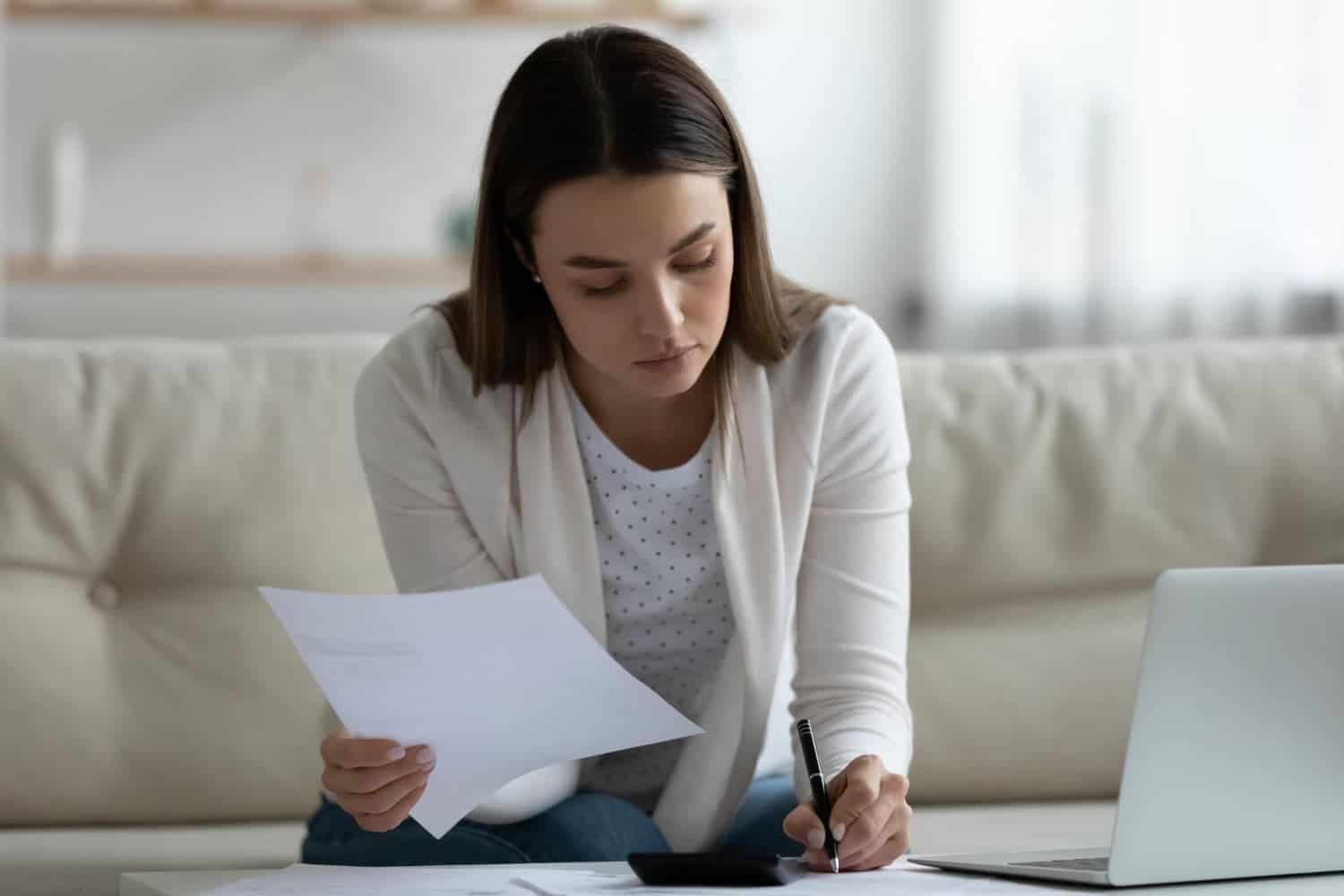 A young woman sits on a couch, holding paperwork and writing notes with a laptop in front of her.