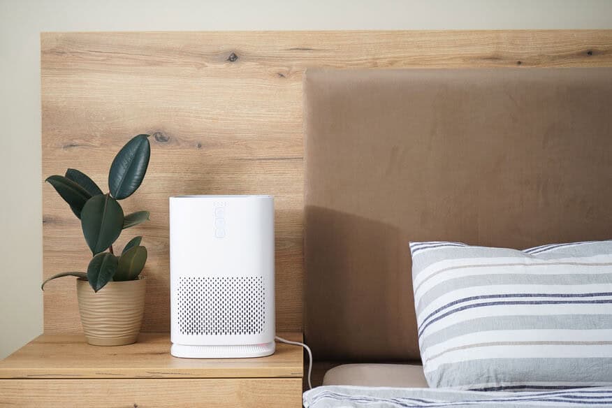 A white air purifier is on a bedside table next to a bed and a plant, creating better hotel air quality in their guest rooms.