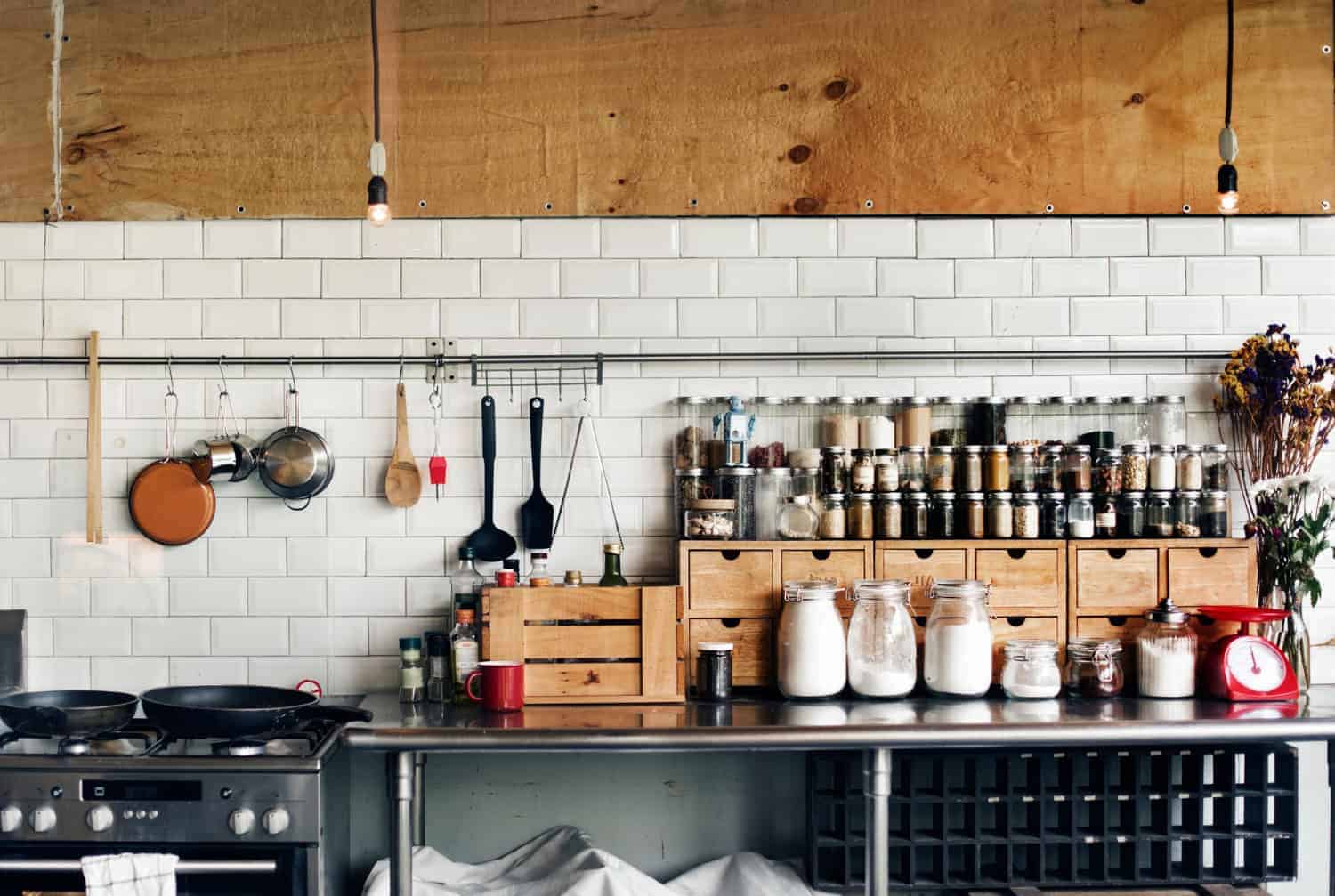 A white tiled wall in a restaurant kitchen is lined with a stovetop, metal prep tables, containers of ingredients and spices, and has kitchen utensils hanging on a wall-mounted bar.