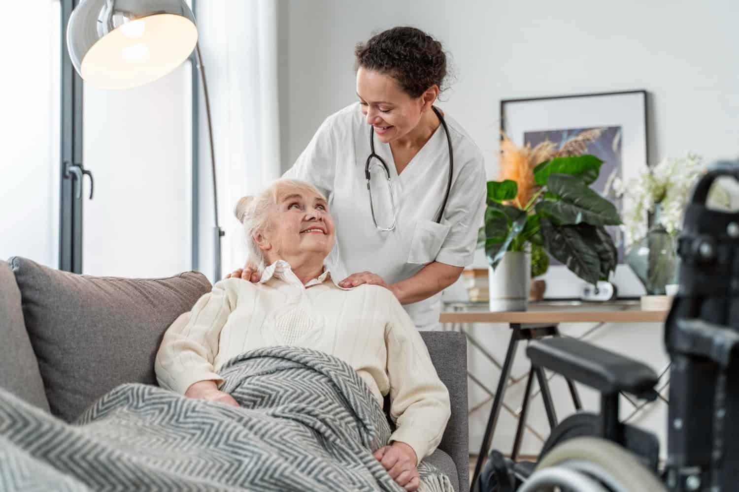 An older woman lays on a couch, with a blanket covering her lower half. A caregiver stands behind her, hands on her shoulders, wearing white scrubs. They're smiling at each other.