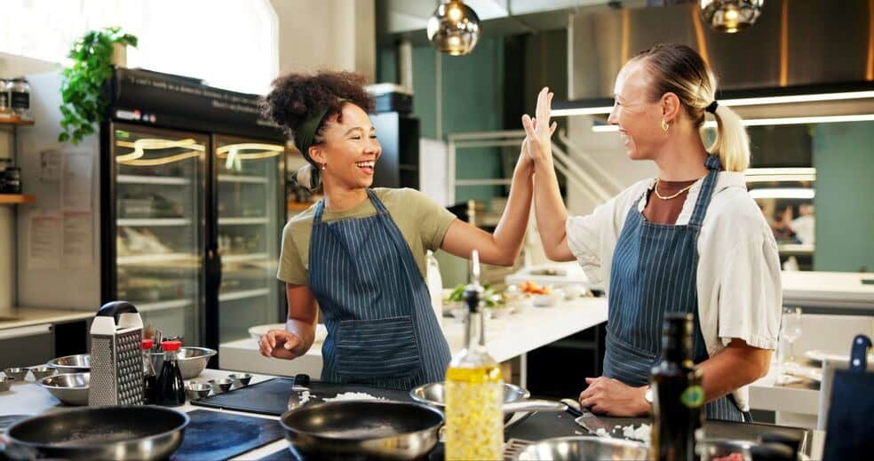 Two female chefs in a commercial kitchen and striped, blue aprons high five each other in celebration of the successful restaurant catering business.