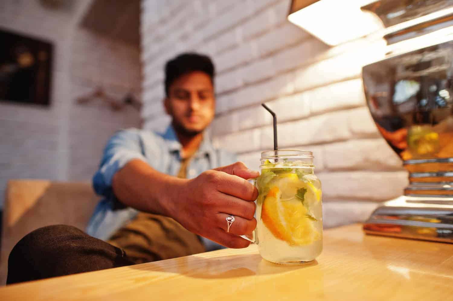 A man sitting at a bar table reaches for a mason jar glass filled with hard seltzer that's garnished with lemon, orange, and lime wedges.