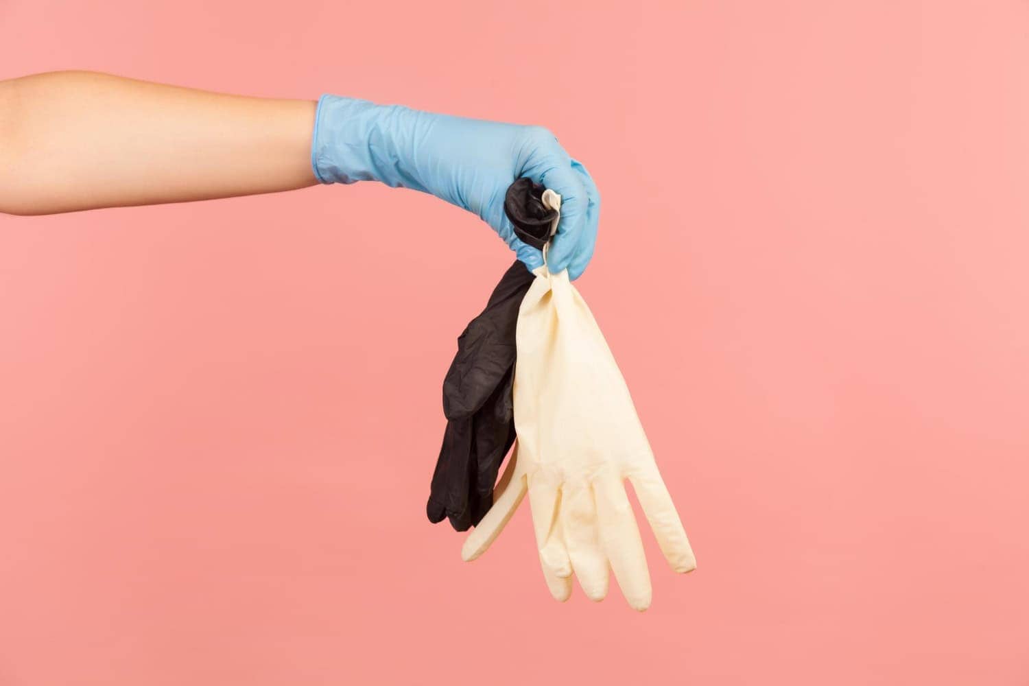 A woman's hand, gloved in a blue latex glove, holds a fistful of white and black food handler gloves against a light pink background.
