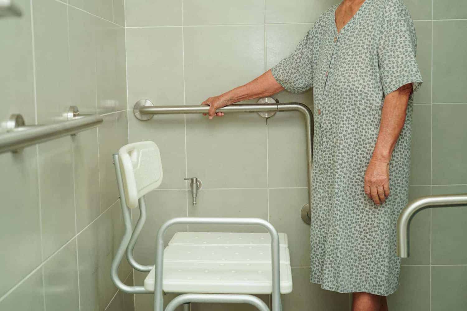 An elderly woman in a nightgown stands inside a large walk-in shower. She's holding on to a wall-mounted metal grab bar, and standing in front of white shower chair.