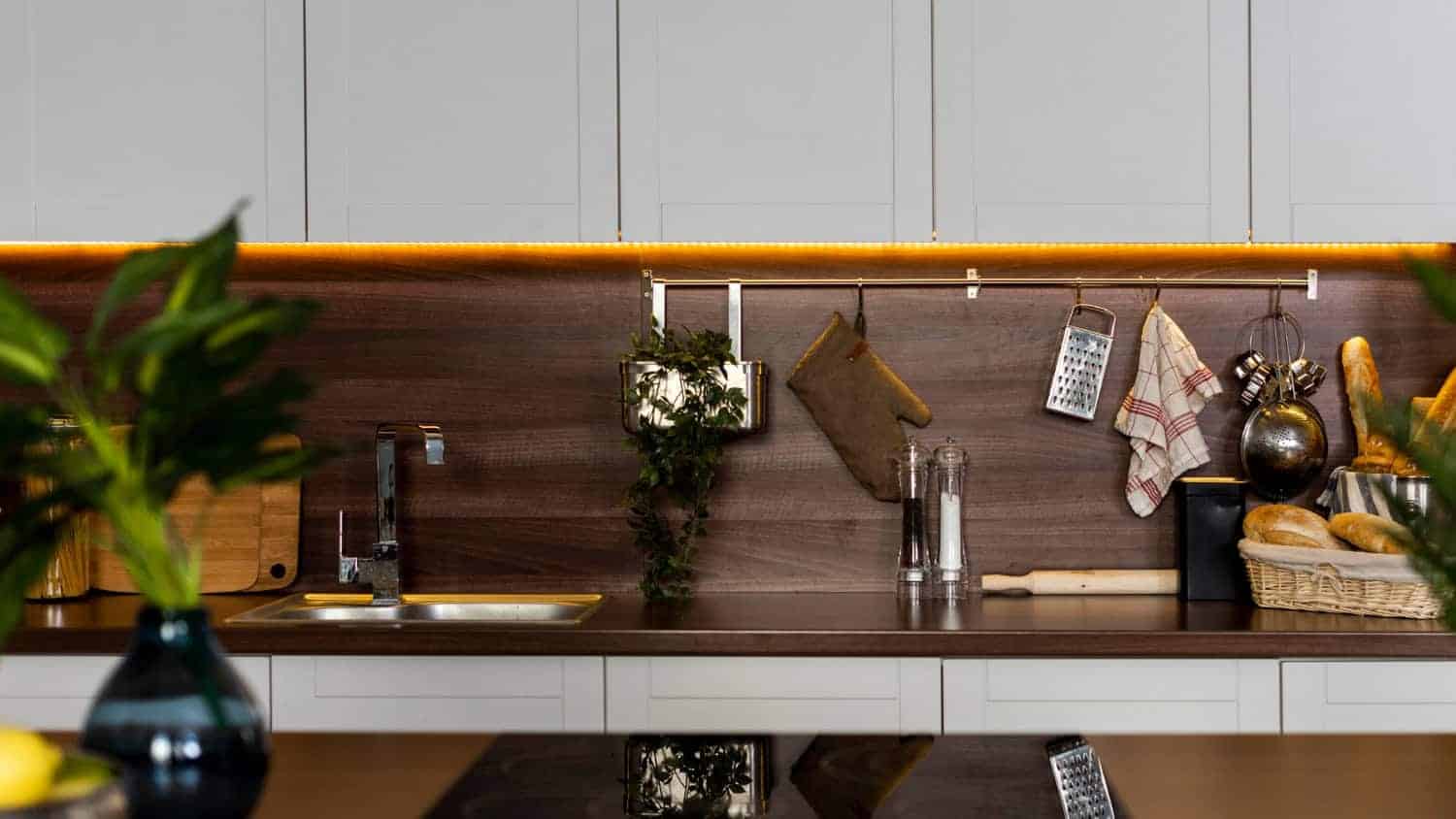 A modern kitchen has white wood cabinets and dark wood countertops and backsplash. The cabinets have soft under cabinet lighting giving off a warm amber glow and illuminating various kitchen accessories.