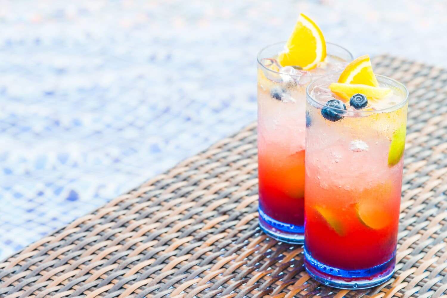 Two glasses are filled with light red liquid and garnished with orange slices, lime slices, mint, and blueberries.