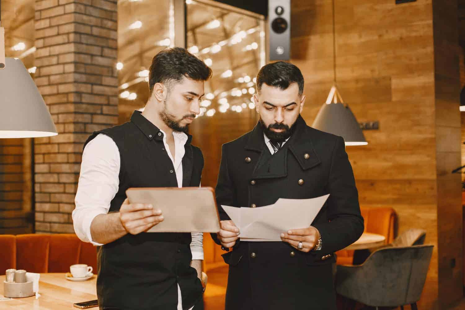 Two waiters study menu pages inside the dining room of a restaurant.