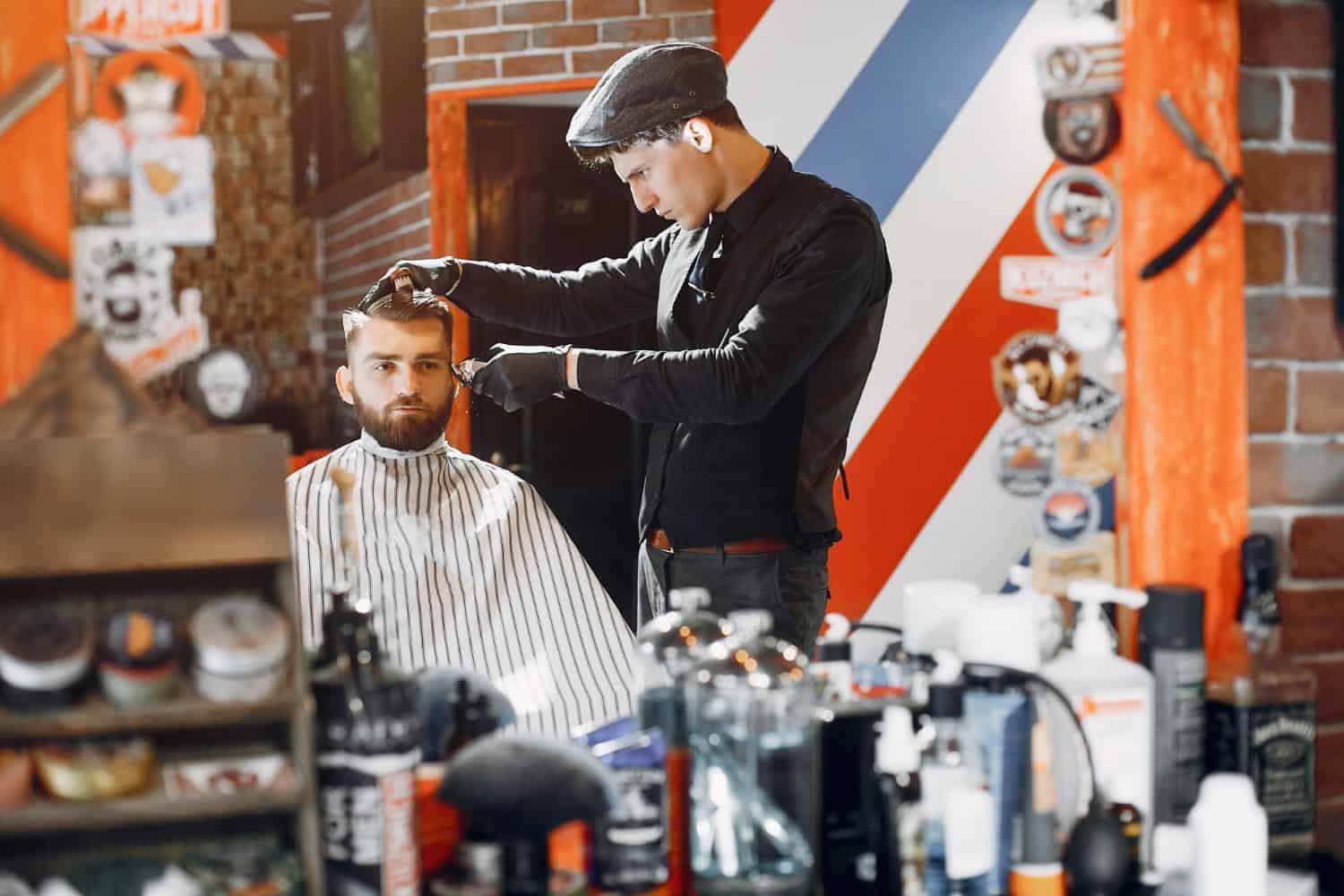 A man receives a hair cut from a male barber in a masculine-decorated salon. In the background, the walls are dark brick and decorated with red, white, and blue striped artwork. The mirror is decorated with stickers and sits behind a counter filled with male hair care and beard products.