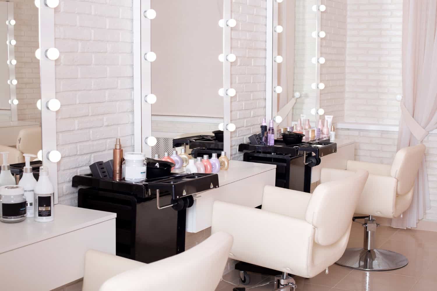 A small salon suite with primarily white decor. Three salon chairs are white leather, in front of three white framed mirrors with vanity lighting hanging on a white brick wall. The salon is bright and clean.