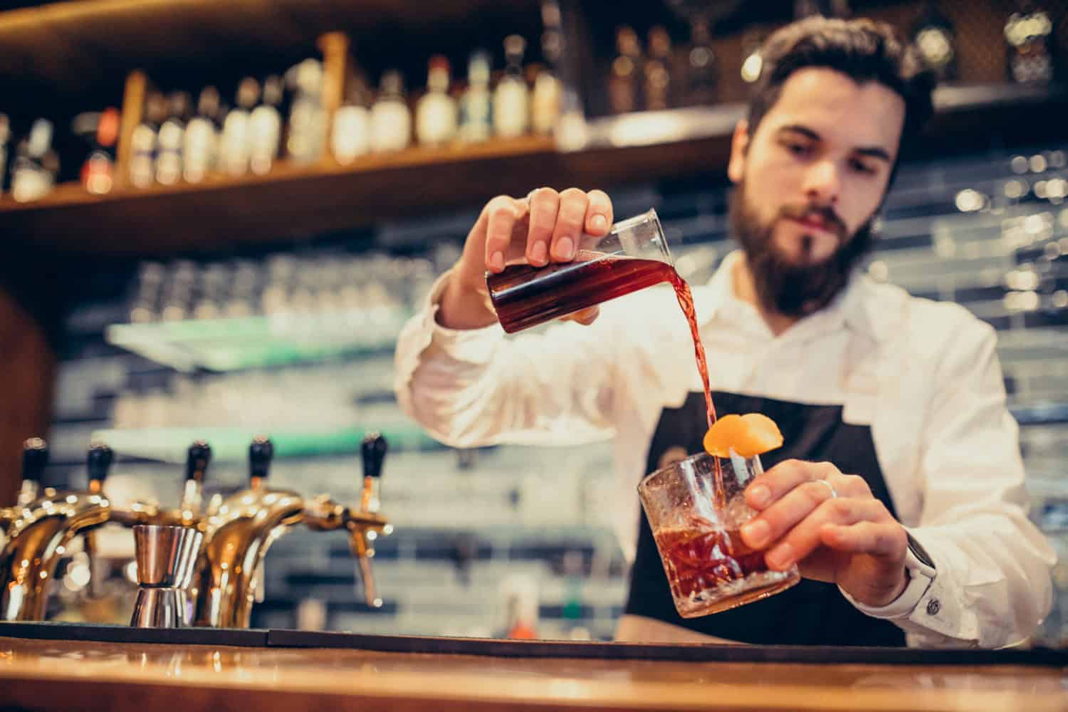 A male bartender creates a craft cocktail, pouring a dark liquid from one glass into a highball glass with ice and an orange slice garnish.