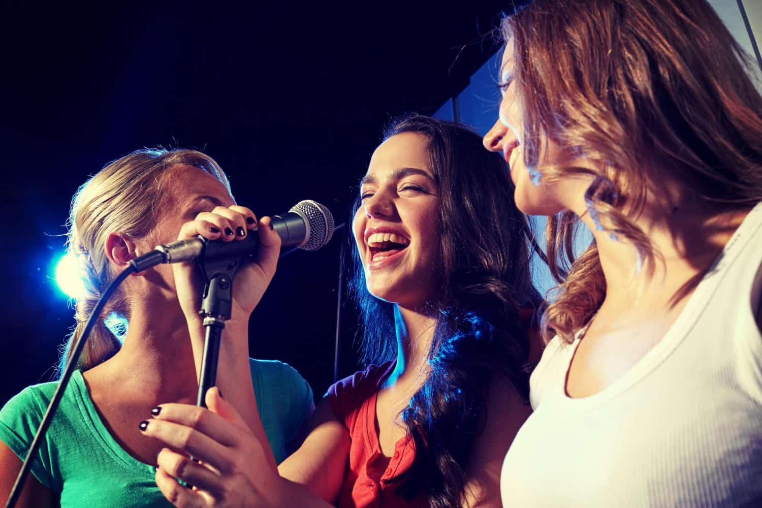 Three women gather around and sing into a microphone during an open mic night at a bar.