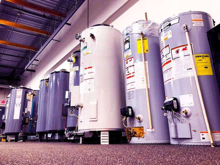 A number of water heaters of various sizes are lined up in a warehouse against a wall.