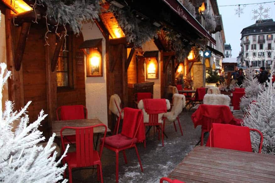 A beautiful wintery outdoor dining area has red chairs and cozy tables. complete with faux fur and blankets.