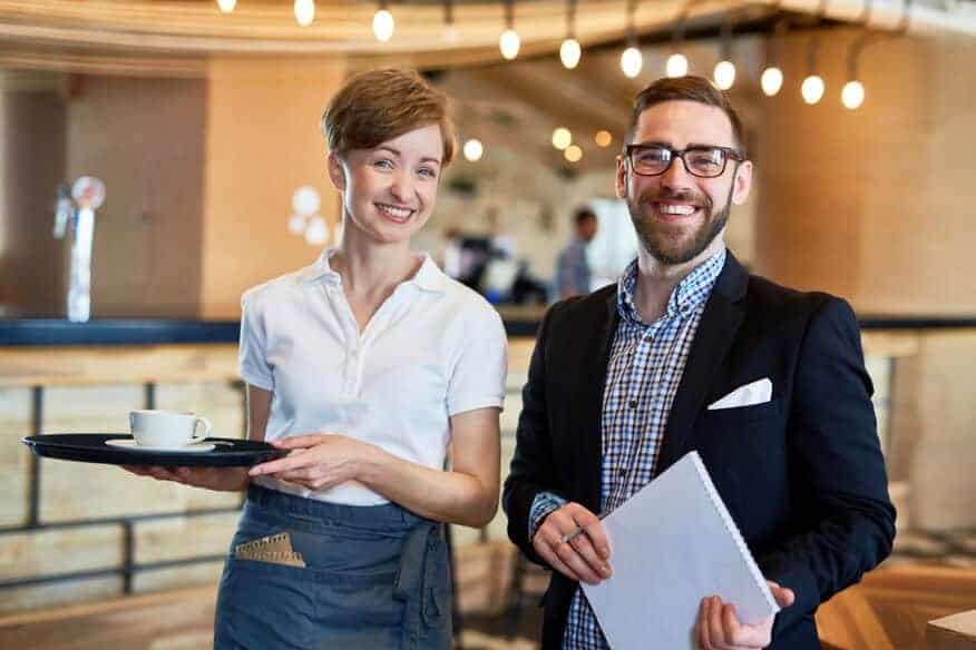 A male restaurant manager and a female server smile while standing in a restaurant. Hiring the right staff is part of learning how to run multiple restaurants.