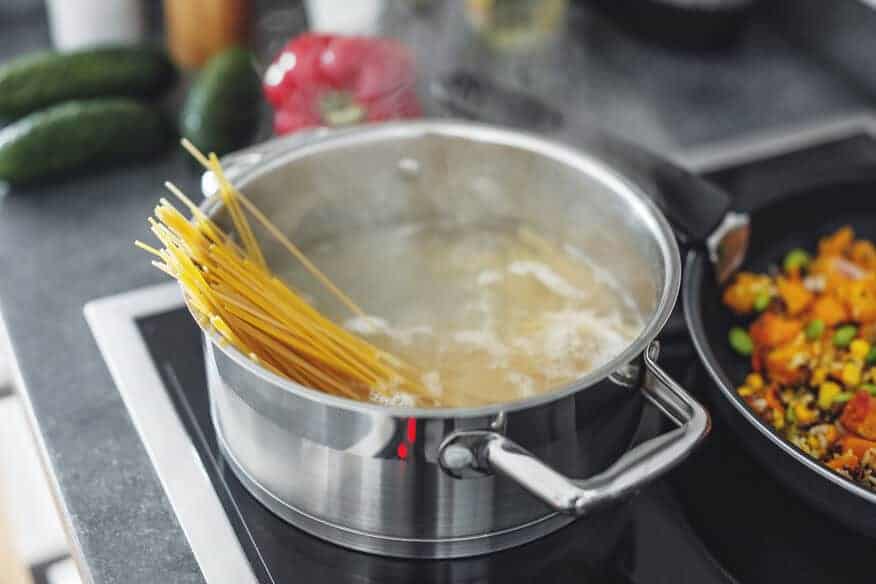 A boiling pot of soft water with hard spaghetti pasta in it is next to a skillet of colorful vegetables.