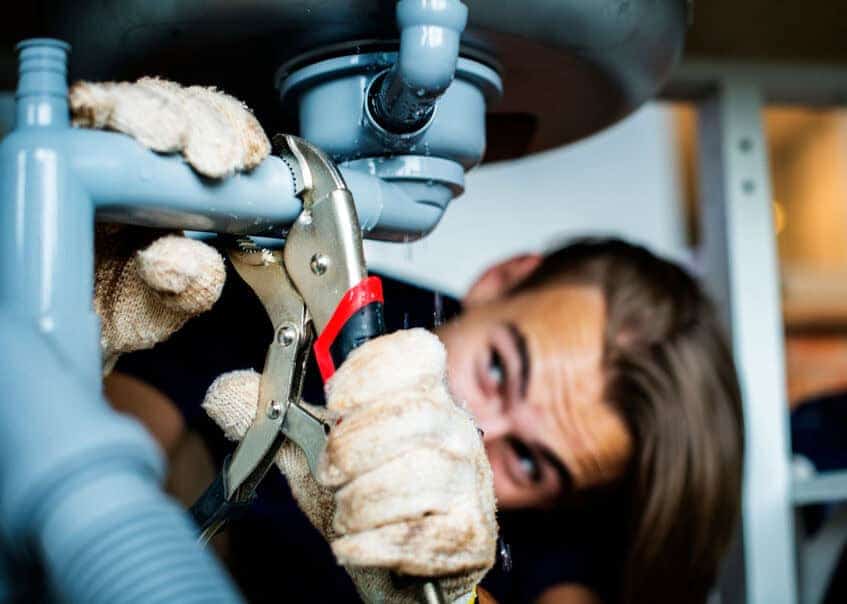 A man attends to salon maintenance by using a wrench to fix the leaking pipes under a sink.