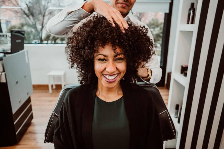 A happy client smiles while the hair stylist adjusts her brown curly hair. Client satisfaction depends on routine salon maintenance.
