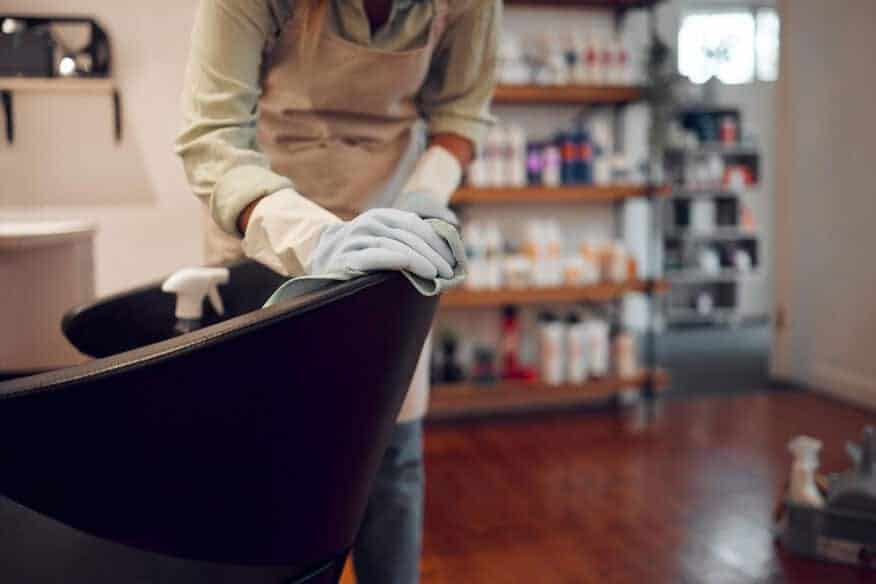 A hair stylist sanitizes and cleans a salon chair using gloves, cleaning solution and hot water as routine salon maintenance.