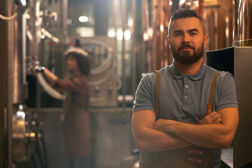 Knowing his brewery boiler repairs are complete, a man stands in front of his brewing equipment with is arms folded and a smile on his face. Behind him, out of focus, a woman performs routine equipment checks. 