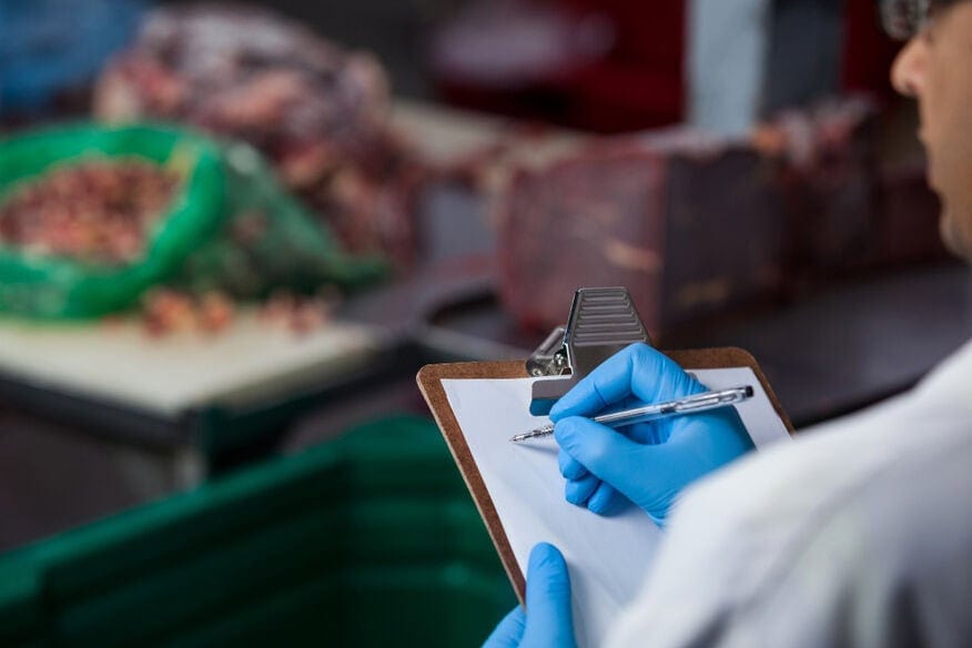 A man documents routine food manufacturing equipment maintenance checks on a clipboard while wearing blue gloves.