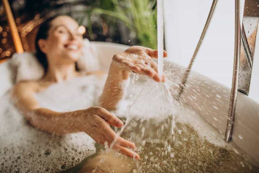A woman in a bubble bath holds her hands under the faucet, enjoying the hot water. She can enjoy her bath because of the hotel's hot water recovery rate.
