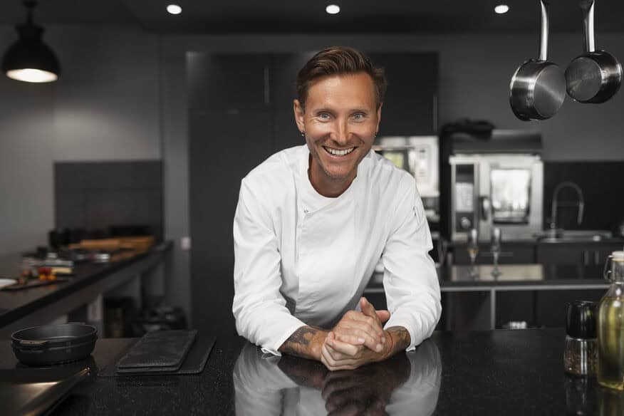A smiling chef leans on a shiny stainless-steel counter, in front of an immaculate kitchen cleaned using hot water.