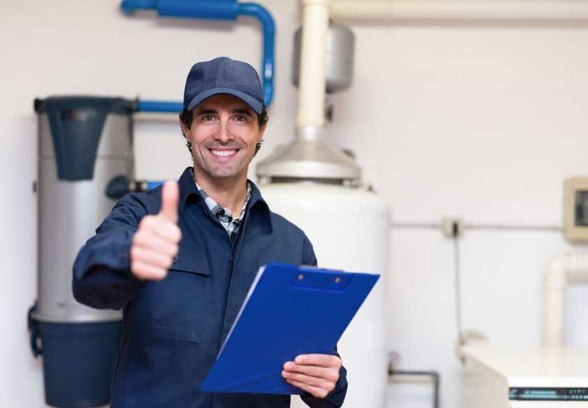A technician in a blue cap and coveralls smiles at the camera. He is giving a thumbs up and holding a blue clipboard, standing in front of water heaters. 