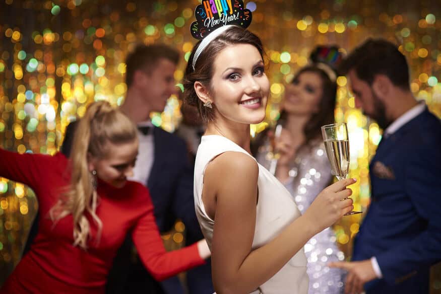 Guests celebrating New Year’s in a hotel ballroom include a smiling woman in a New Year's Eve hat holding champagne in front of dancing and a gold curtain. The event may not matter, but the need to address hotel event space hot water demand during peak events certainly warrants attention. 