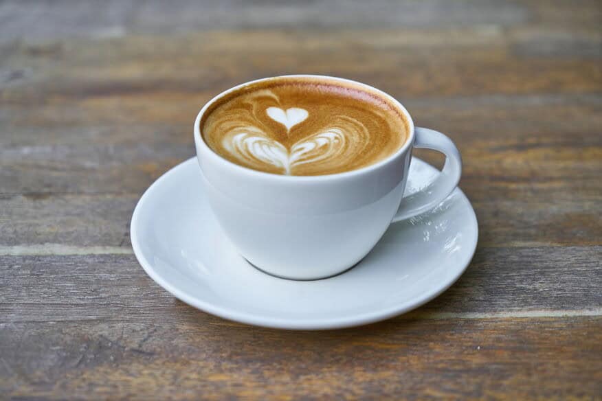 A close picture of a white mug and saucer for a fresh latte coffee, with two foam hearts. Coffee and beverage tastes suffer from poor hotel water quality. 