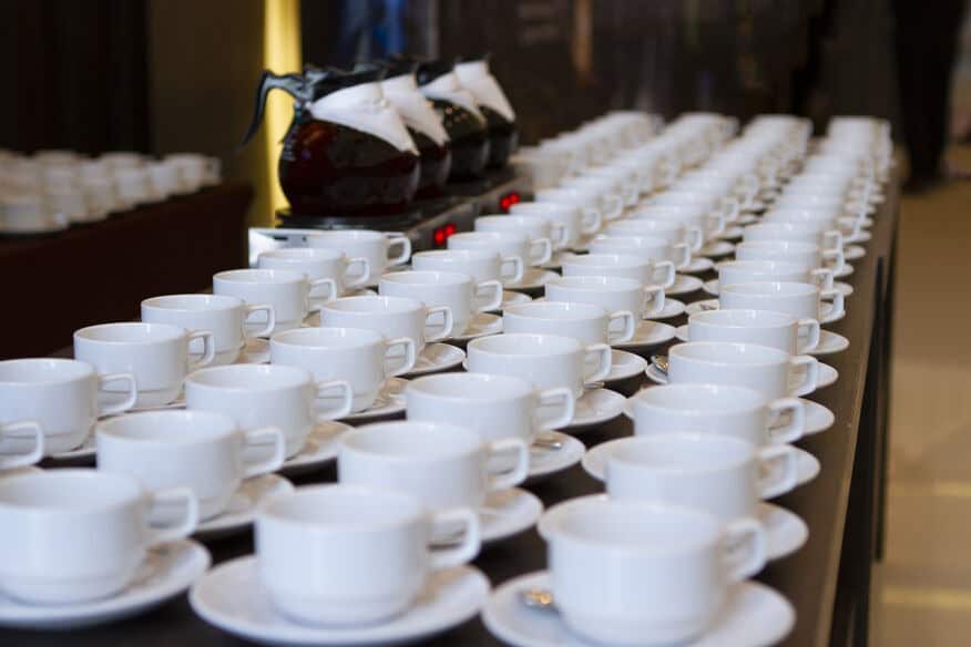 A conference beverage station with filled coffee carafes and lines of empty coffee cups demonstrates hotel event space hot water demand during peak service.