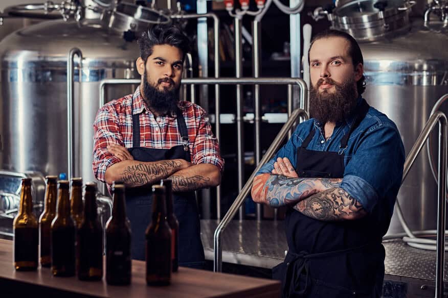 Two hipster men with beards and arm tattoos stand in front of their beer tanks with their arms crossed. The hot water demand in a taproom and on the production floor can vary greatly.