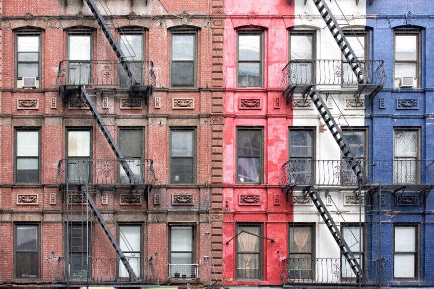 An old New York apartment building, with brown, red, white, and blue exteriors, and fire escapes crawling up the outside wall.