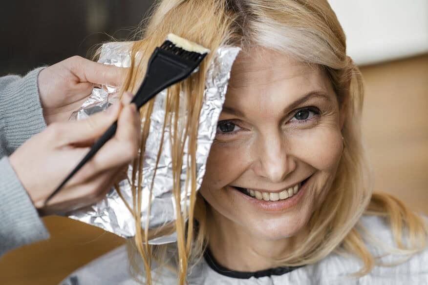 A smiling woman gets her gray hair colored using foils in a salon.