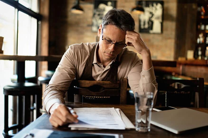 A distraught restaurant owner sits in the dining room with his hand on his head, while going through the costs of repairing a commercial water heater. 