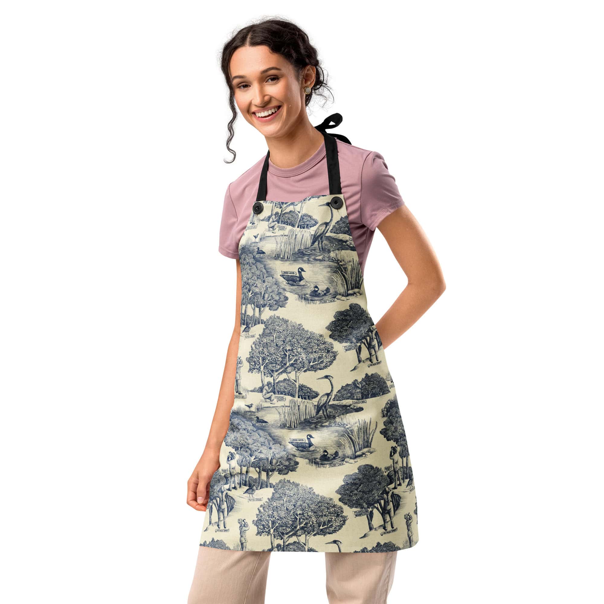 Woman wearing the Birds & Birders Toile De Jouy apron over a pink top, standing with hand on hip.