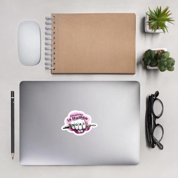 Lay flat desktop scene with eyeglasses, pen, computer mouse, notepad and a laptop with the No AI sticker affixed to the top.