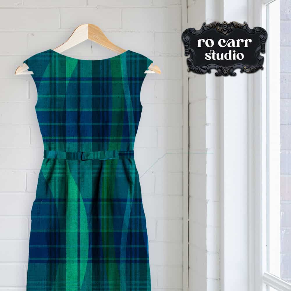 A sleeveless belted shift dress in teal and navy tartan, hanging near a window.