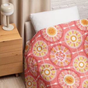 Tour de Citrus comforter on a bed with a white pillow beside a wooden nightstand, showing the coral and golden yellow hand-drawn sun medallion print.