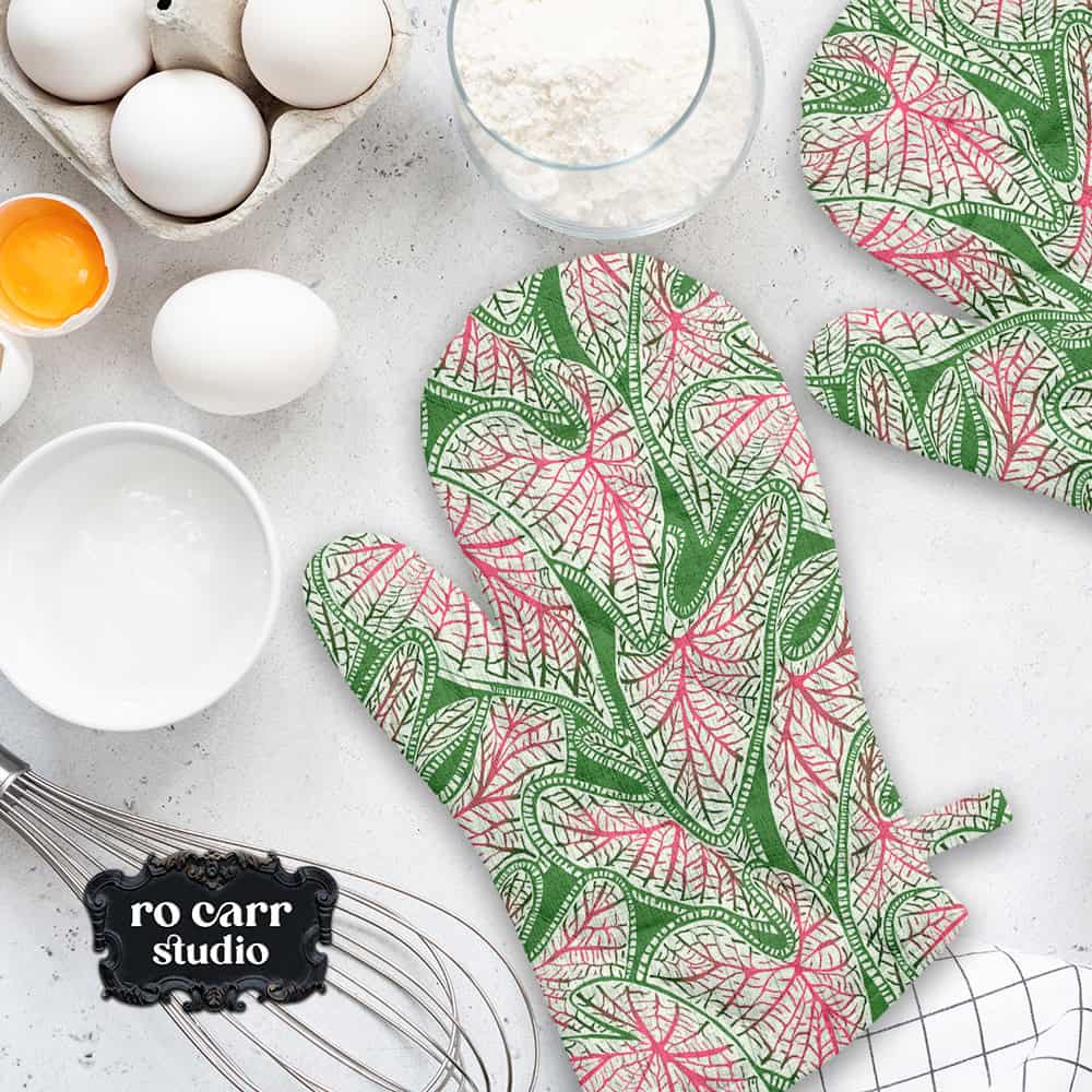Caladium print oven mitts on a baking counter beside eggs, flour, and a whisk.