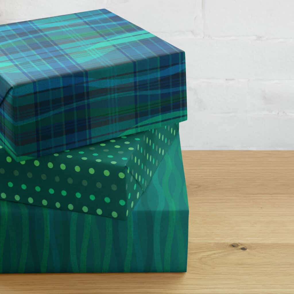 Three gifts wrapped in the Breaking Plaid wrapping paper set and stacked on a wooden table: the teal, blue, and green plaid broken by flowing wave shapes on top, a dark green polka dot coordinate in the middle, and a green wave coordinate on the bottom.