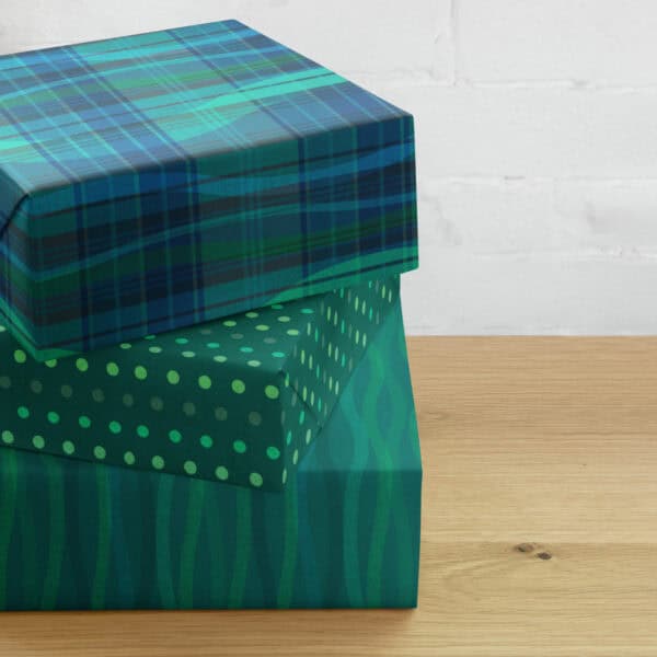 Three gifts wrapped in the Breaking Plaid wrapping paper set and stacked on a wooden table: the teal, blue, and green plaid broken by flowing wave shapes on top, a dark green polka dot coordinate in the middle, and a green wave coordinate on the bottom.