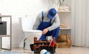 A friendly Service Champions plumber in blue uniform carefully inspects a bathroom toilet, ready with tools to ensure your comfort.