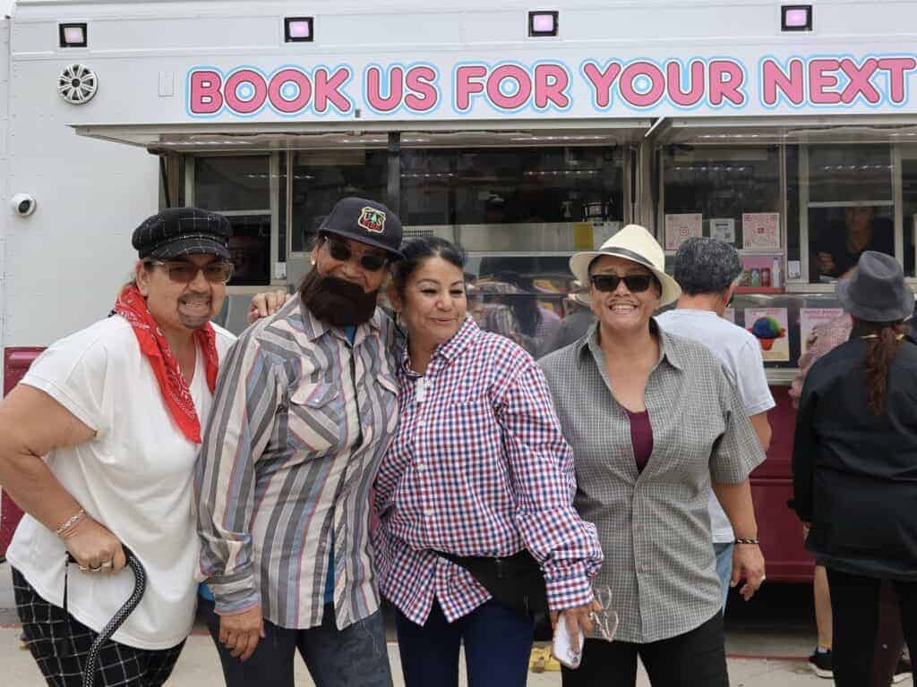 Four adults, dressed casually and wearing hats, stand smiling in front of a food truck displaying a “Book Us For Your Next” sign, enjoying the friendly community atmosphere in Anaheim. In the background, two individuals wait eagerly at the truck window for complimentary ice cream served by Service Champions—Southern California’s trusted local experts dedicated to dependable HVAC, plumbing, and drain services. Their presence reflects a commitment to helpfulness and customer-focused care throughout the community.