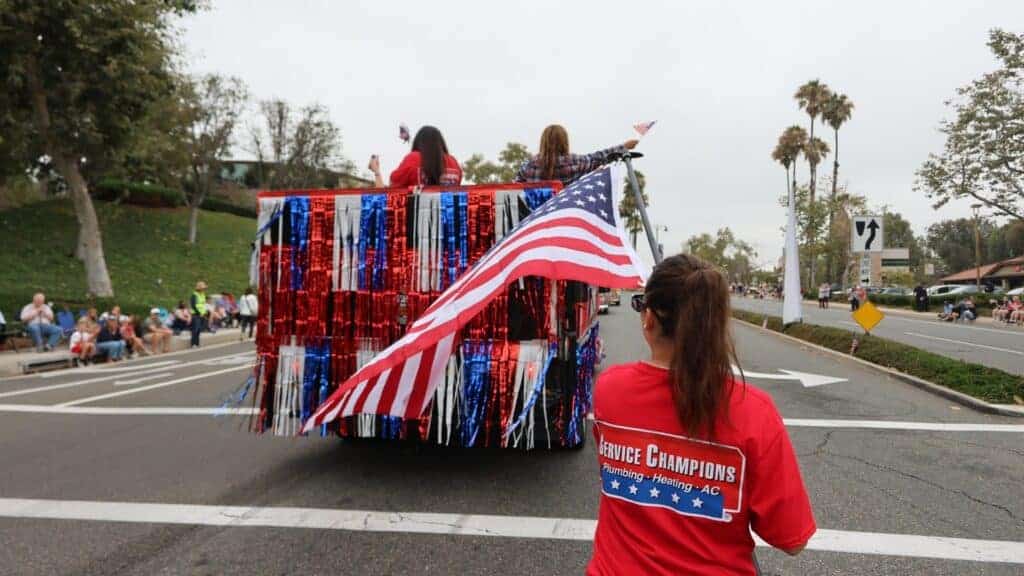 Service Champions team members, wearing vibrant red shirts, proudly walk behind an America 250 Parade float adorned with patriotic red, white, and blue streamers and an American flag along a city street in Mission Viejo. The festive scene celebrates Service Champions’ trusted presence as Southern California’s local HVAC, plumbing, and drain professionals—dedicated to providing dependable home comfort solutions while supporting their community.